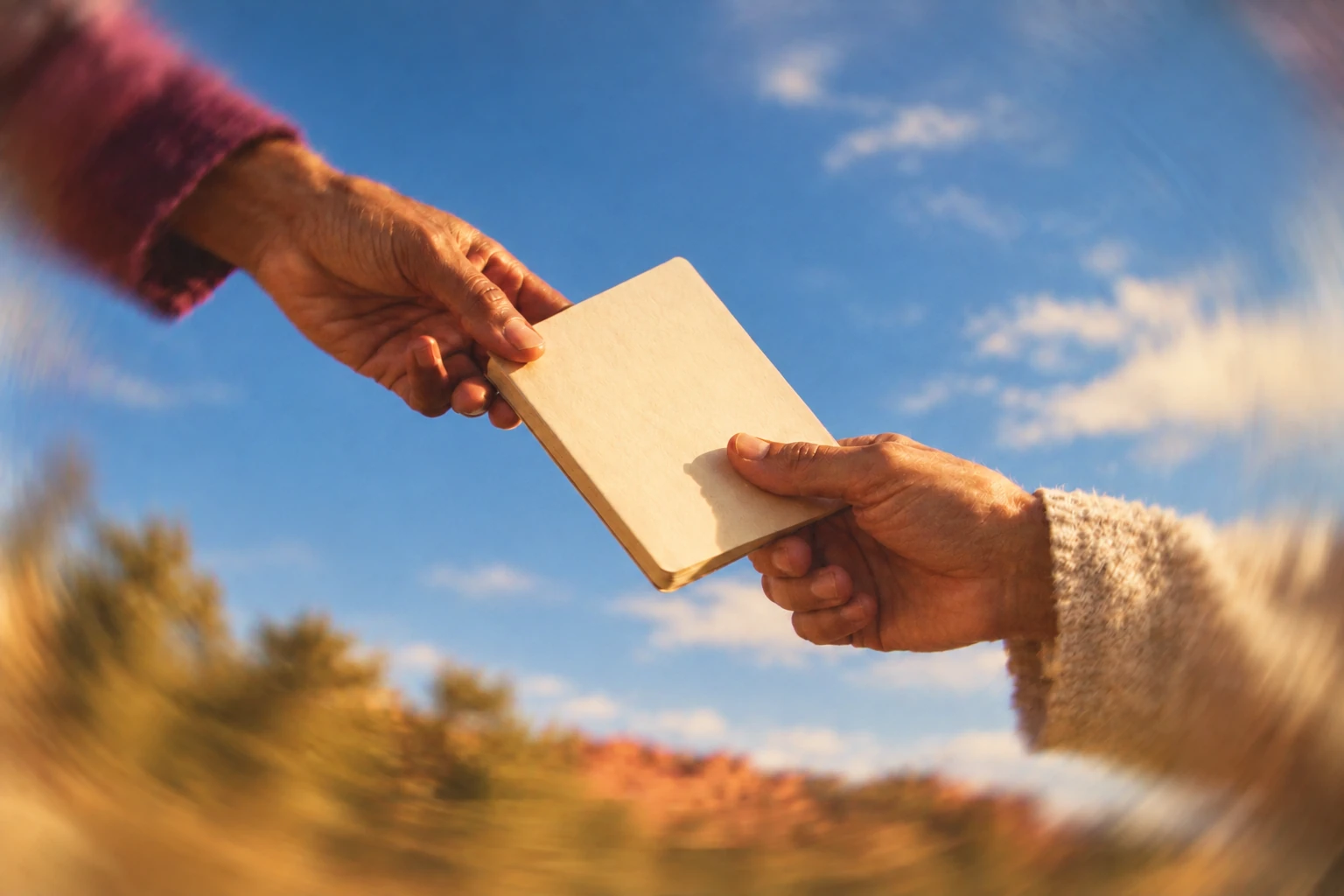 Two hands pass a closed cream notebook against blue sky.
